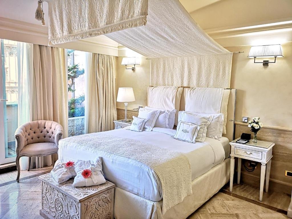 quarto do Hotel Barocco, um dos hotéis no centro de Roma, com cama de casal grabde, mesinha e luminária de ambos os lados, um dossel que passa por cima da cama, tudo em tons de bege clarinho e branco, há janela com vista com cortinas claras e poltrona