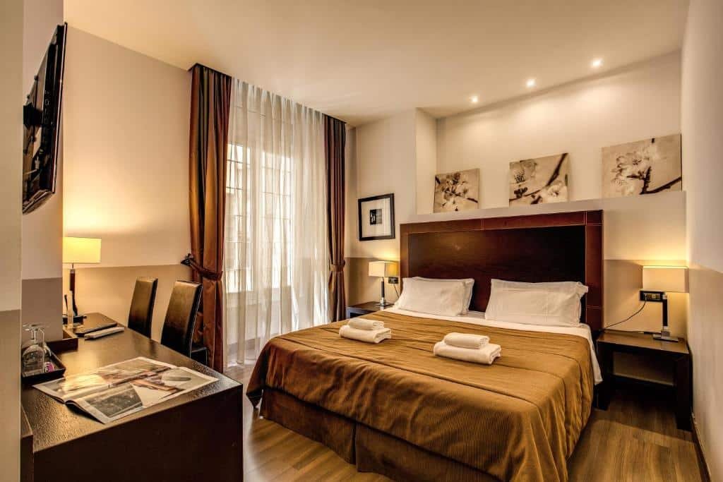 quarto do Hotel Boutique Nazionale, um dos hotéis boutique em Roma, com cama de casal grande, mesinha e luminárias de ambos os lados, quadros na parede e detalhes em tons de marrom na cama e nas cortinas de janelas grandes, há uma tv, mesa e cadeira em frente