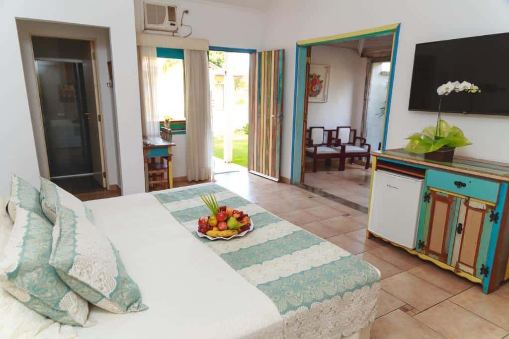 Quarto com cama, bandeja de frutas e detalhes em turquesa no Hotel Fazenda Hípica Atibaia. Há portas abertas que levam a uma sala de estar e ao exterior. Também há uma TV montada na parede.