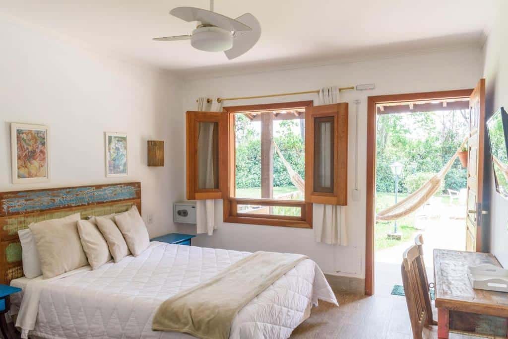 Um quarto bem iluminado do Hotel Recanto da Cachoeira com uma cama de casal, janela e porta abertas, ventilador de teto, obras de arte, escrivaninha e rede do lado de fora.