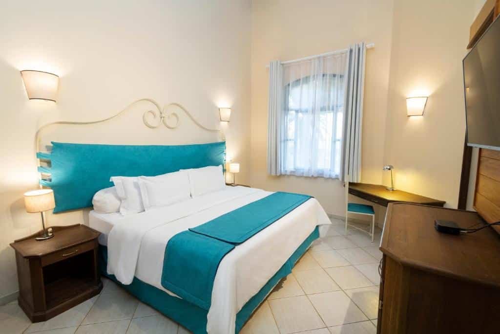 A imagem mostra um quarto do Hotel Villa Rossa com uma cama de casal grande, coberta com lençóis brancos e uma colcha azul turquesa, assim como uma cabeceira estofada em azul combinando. As paredes são de cor clara, e o quarto tem iluminação suave com luminárias montadas nas paredes, criando um ambiente aconchegante. Há uma pequena mesa de trabalho com uma cadeira próxima à janela, que está coberta por uma cortina branca. Ao lado da cama, há um criado-mudo de madeira com um abajur, e na frente da cama, uma televisão montada na parede.