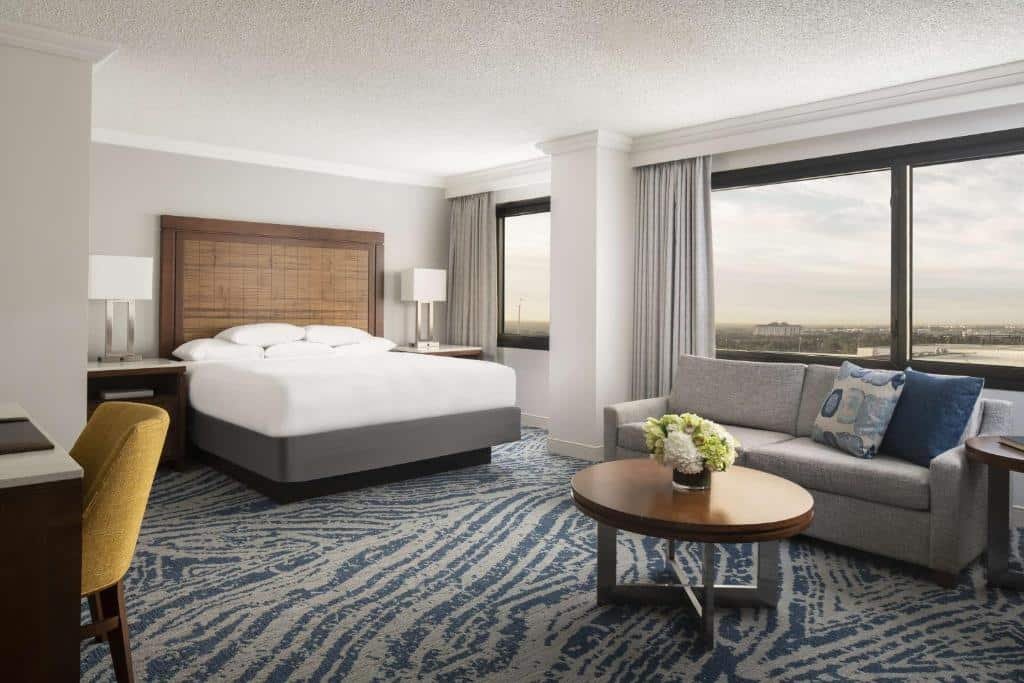 quarto do Hyatt Regency Orlando com cama de casal, mesinha e luminárias em ambos os lados, em frente há uma mesinha com sofá e, do outro lado, duas janelas com cortinas e em frente com balcão e cadeira