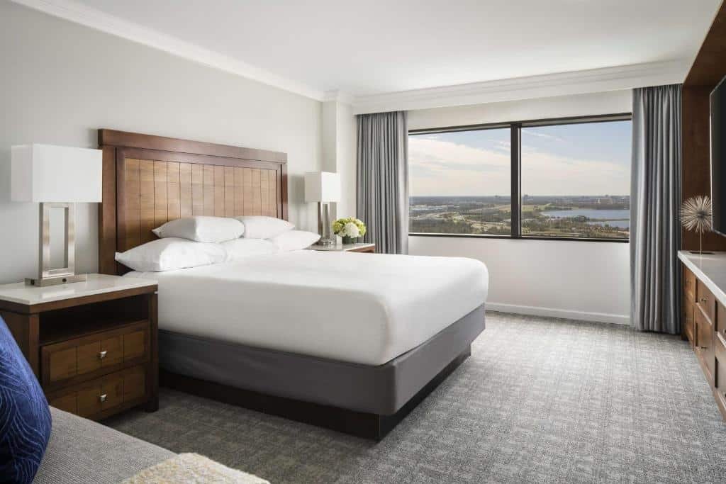 quarto amplo com cama de casal com mesinha e luminária de ambos os lados com janela grande de vidro e cortinas cinza, há ainda um sofá e tv em frente, no Hyatt Regency Orlando, uma das opções de onde ficar em Orlando