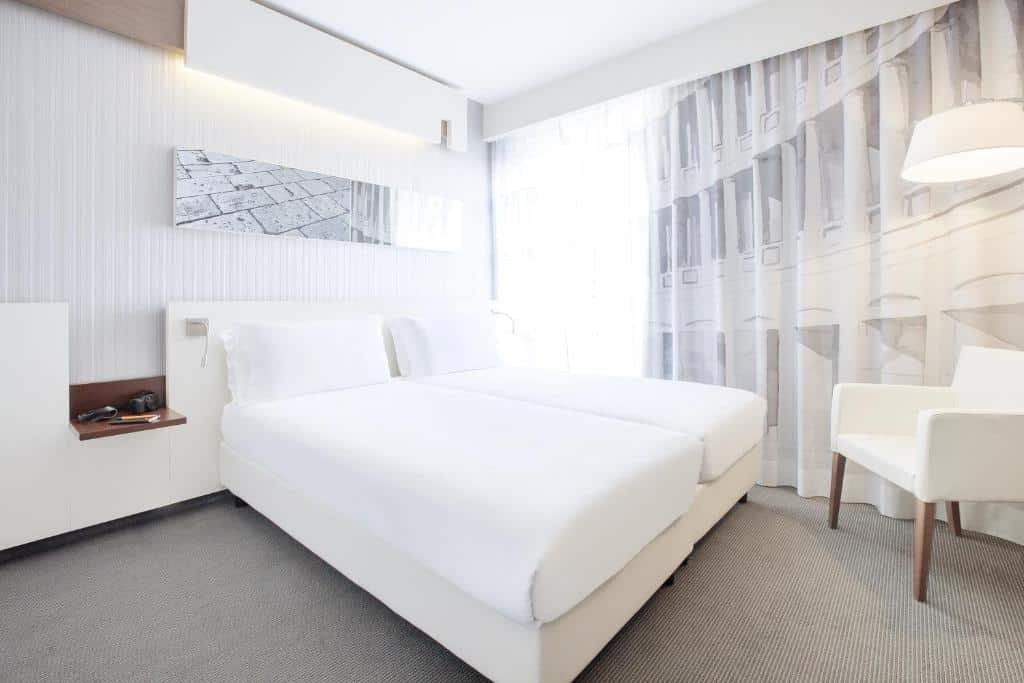 quarto espaçoso do Ibis Styles Roma Eur, com decoração toda em branco e com cama de casal, mesinha de cabeceira, poltrona, bancada e cortinas brancas na janela grande