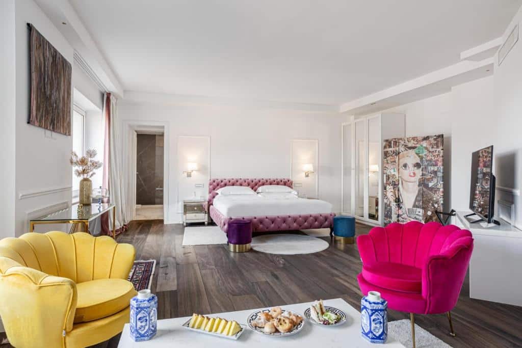 quarto amplo do Monastero dei Santi, um dos hotéis boutique em Roma, com cama de casal, mesinha e luminária de ambos os lados, duas poltronas com encosto de concha rosa e amarelo, mesa de centro com café da manhã servido, há obras de arte no quarto e a janela é grande, tudo em tons brancos e leves