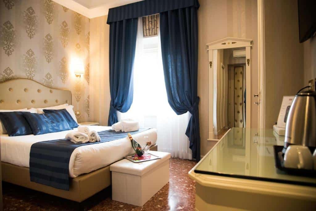 quarto do Navona Queen Rooftop, um dos melhores hotéis em Roma, com cama de casal, baú, janela grande com cortinas azuis combinando com detalhes da cama, mesinha e luminária de ambos os lados, mesa à frente com chaleira