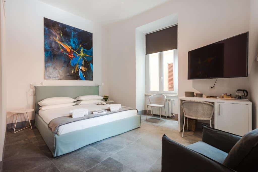 quarto do Nikis Collection Navona, um dos hotéis no centro de Roma, com cama bem grande, janela comprida com persiana, tv com mesa e cadeira, há uma poltrona em frente, um quadro abstrato colorida acima da cama, que conta ainda com mesinha de ambos os lados