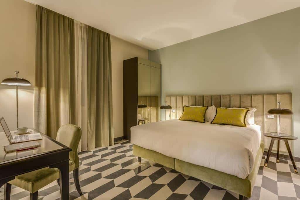 quarto do Otivm Hotel, um dos melhores hotéis em Roma, com cama de casal bem grande com luminária e mesinha de ambos os lados, o chão é xadrez e as janelas possuem cortinas pesadas, em tons de verde para combinar com as fronhas, há ainda uma cadeira e mesa com tv à frente