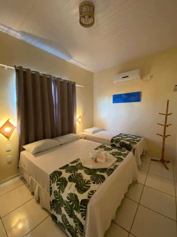 quarto do Pousada Do Porto com cama de casal e uma de solteiro com uma mesinha no meio delas, há luminárias de parede baixas e uma janela com cortinas escuras atrás, do outro lado há um ar-condicionado e um mancebo