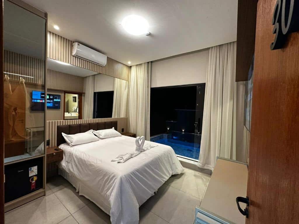 A imagem mostra o interior de um quarto da Pousada Villas em Bonito, Mato Grosso do Sul. O quarto é decorado de maneira moderna e aconchegante, com tons neutros e detalhes em madeira. A cama de casal é coberta com roupas de cama brancas impecáveis e há uma toalha dobrada em forma de cisne sobre a cama.

À esquerda, há um armário com portas de vidro espelhado e um frigobar embutido. Acima do frigobar, há um suporte para malas. Na parede oposta à cama, há uma televisão de tela plana montada, permitindo entretenimento para os hóspedes. O quarto possui ar-condicionado montado na parede para maior conforto.

Uma grande janela com cortinas claras oferece uma vista para a área externa, onde se pode ver uma piscina privativa iluminada à noite, proporcionando um ambiente relaxante e luxuoso.