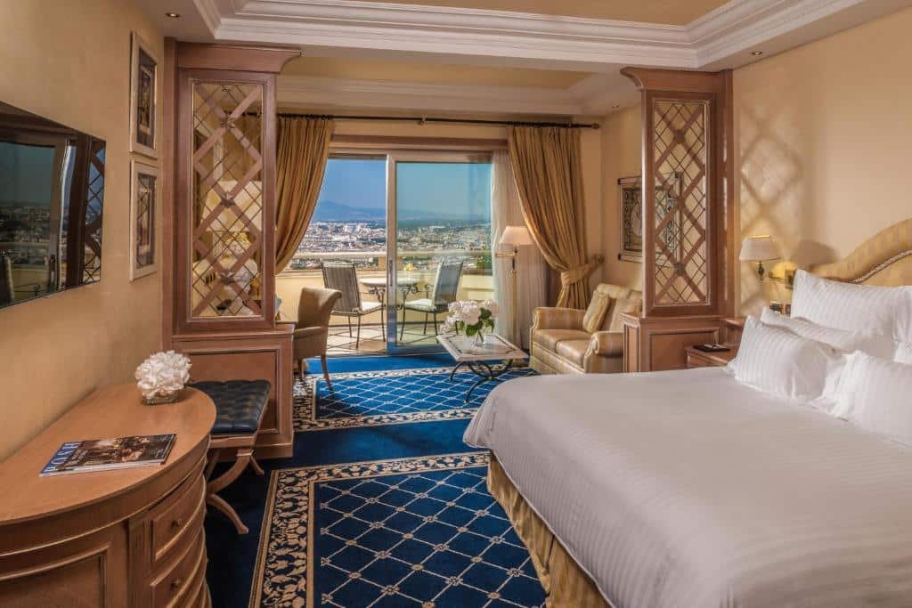 quarto do Rome Cavalieri, A Waldorf Astoria Hotel, um dos hotéis de luxo em Roma, com cama de casal grande, cômoda e mesa e TV à frente, há uma área de estar com sofá e uma sacada com mesa e cadeiras com vista da cidade, tudo em tons claros e detalhes em azul