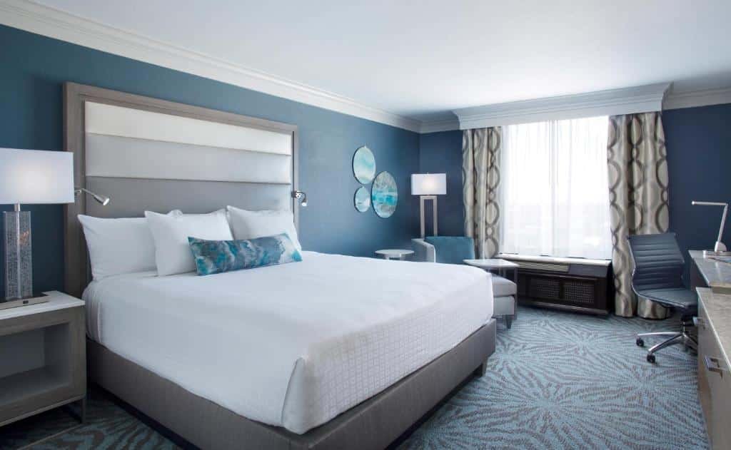 quarto do Rosen Centre Hotel Orlando Convention Center com janela de cortinas claras, sofa abaixo com uma luminária de chão, mesinha, poltrona e cama de casal