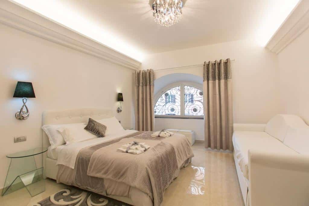 quarto do San Pietro Grand Suite, um dos melhores hotéis em Roma, com cama de casal, mesinha e luminária de ambos os lados, sofá à frente e janela arredondada em cima com janelas bege combinando com os outros detalhes do quarto