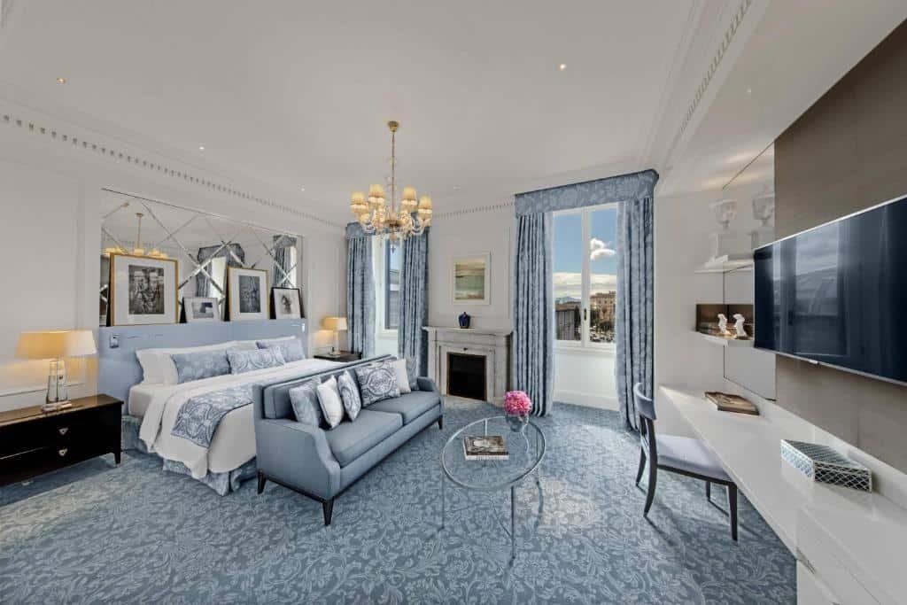 quarto do The St. Regis Rome, um dos hotéis de luxo em Roma, com espaços amplos em detalhes em azul, com cama bem grande, lustre dourado, mesinha de centro com sofá e mesa e poltronas, e duas janelas grandes com vista da Praça da República