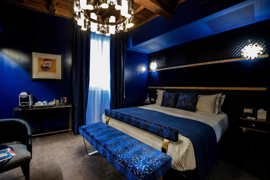quarto do Torre Argentina Relais - Residenze di Charme, um dos hotéis boutique em Roma, com cama de casal grande, lustre redondo e trabalhado, quadros na parede, mesa com cadeira de madeira e todos os detalhes em um azul escuro luxuoso, há mesinha e luminária de ambos os lados da cama e a janela é comprida e grande