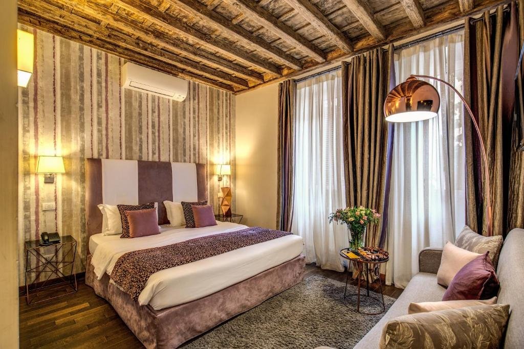 quarto do Trevi Beau Boutique Hotel, com cama de casal grande em detalhes de madeira e lilás, as janelas são grandes e há cortinas, há um sofá em frente, mesinha e luminária de ambos os lados e ar-condicionado, tudo bastante detalhado