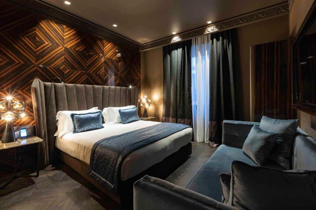 quarto do U-Visionary Roma Hotel, um dos hotéis no centro de Roma, com cama de casal, mesinha e luminária de ambos os lados, há uma tv e sofá em frente, do outro lado, uma janela comprida com mais luzinhas pelo teto, as cores são sóbrias, variando entre branco, azul e marrom