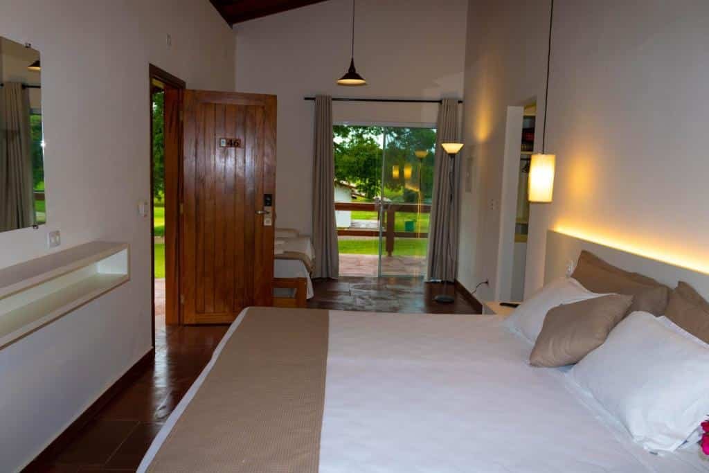 Quarto do Vale Das Águas Fazenda Resort com cama grande, paredes brancas e decoração em madeira. O quarto tem uma sacada com vista para uma área gramada.