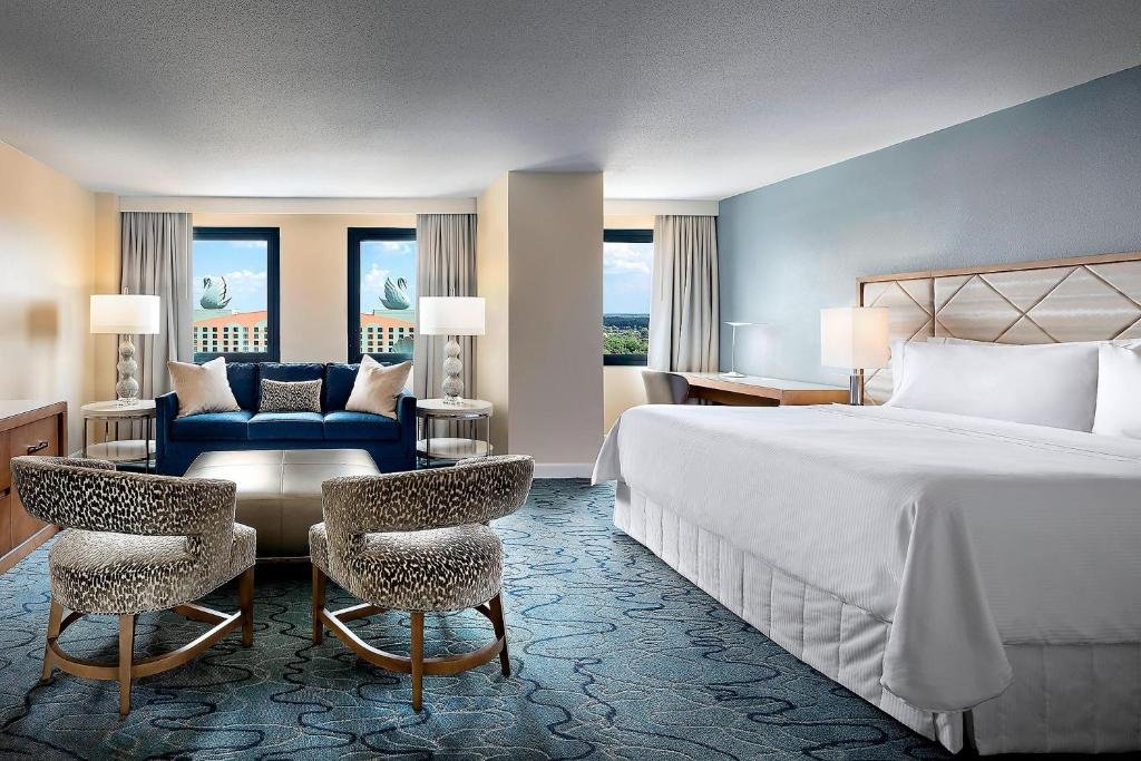 quarto grande do Walt Disney World Dolphin com mesa e cadeiras grandes em frente a um sofá, há uma divisão para uma bacana com cadeira de trabalho e uma cama grande com mesinha e luminária de ambos os lados, também há janelas e o ambiente é claro