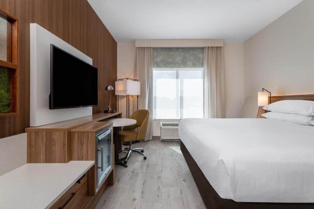 quarto do Wyndham Garden Orlando Universal / I Drive com tv e balcão com frigobar e uma cadeira em frente à cama de casal, no outro lado, uma janela grande com cortinas e luminária de chão