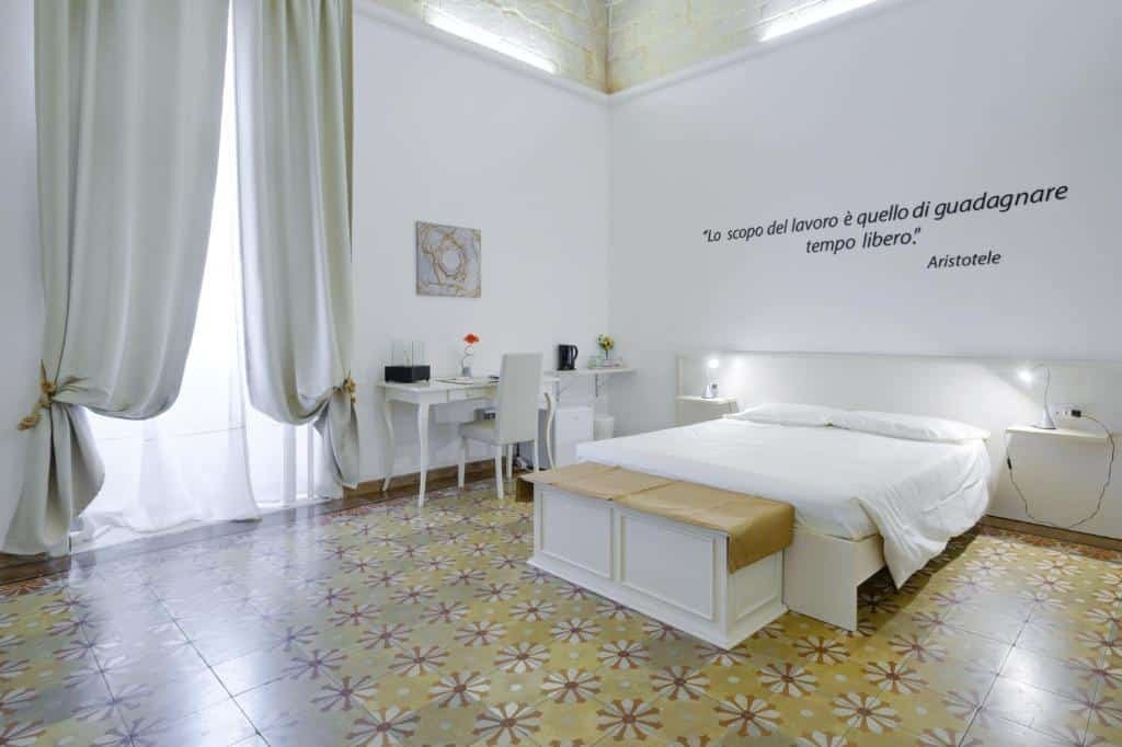 Quarto do Ada Suites & Spa com uma cama de casal bem ao centro e um baú aos pés da cama. Além disso, o piso do quarto de azulejos frios, as paredes são brancas com uma escrita em cima da cama e do lado esquerdo da imagem tem uma escrivaninha com uma cadeira, um quadro na parede e uma porta coberta pela cortina.