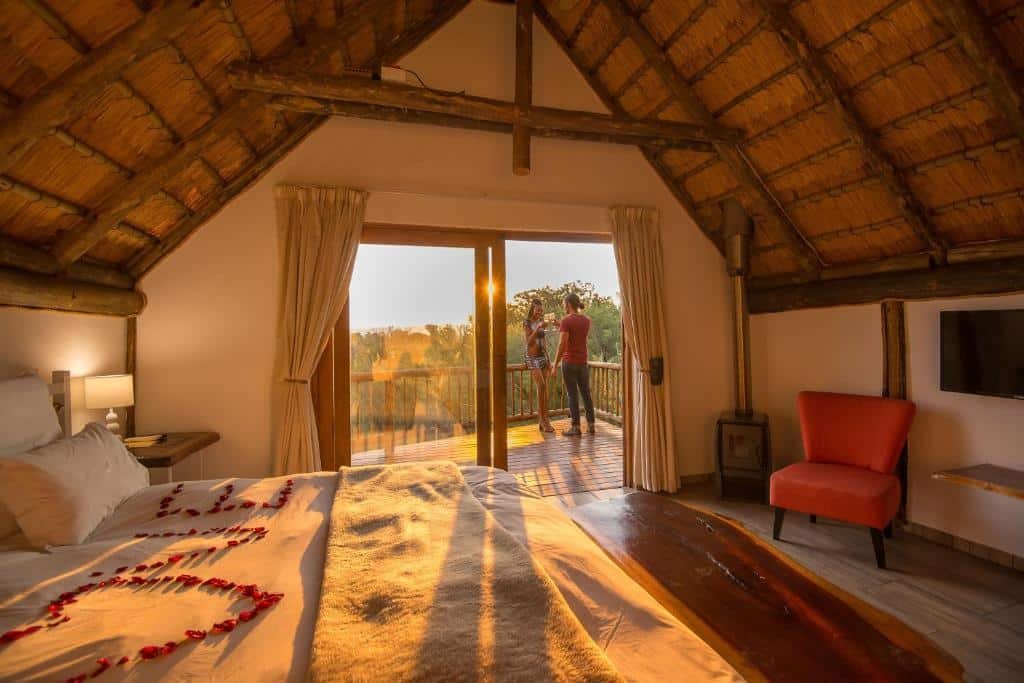 Quarto aconchegante do  Amadwala Lodge com telhado de palha, cama com pétalas de rosas, uma cadeira vermelha e um casal na varanda com vista para o pôr do sol. Representa Joanesburgo.
