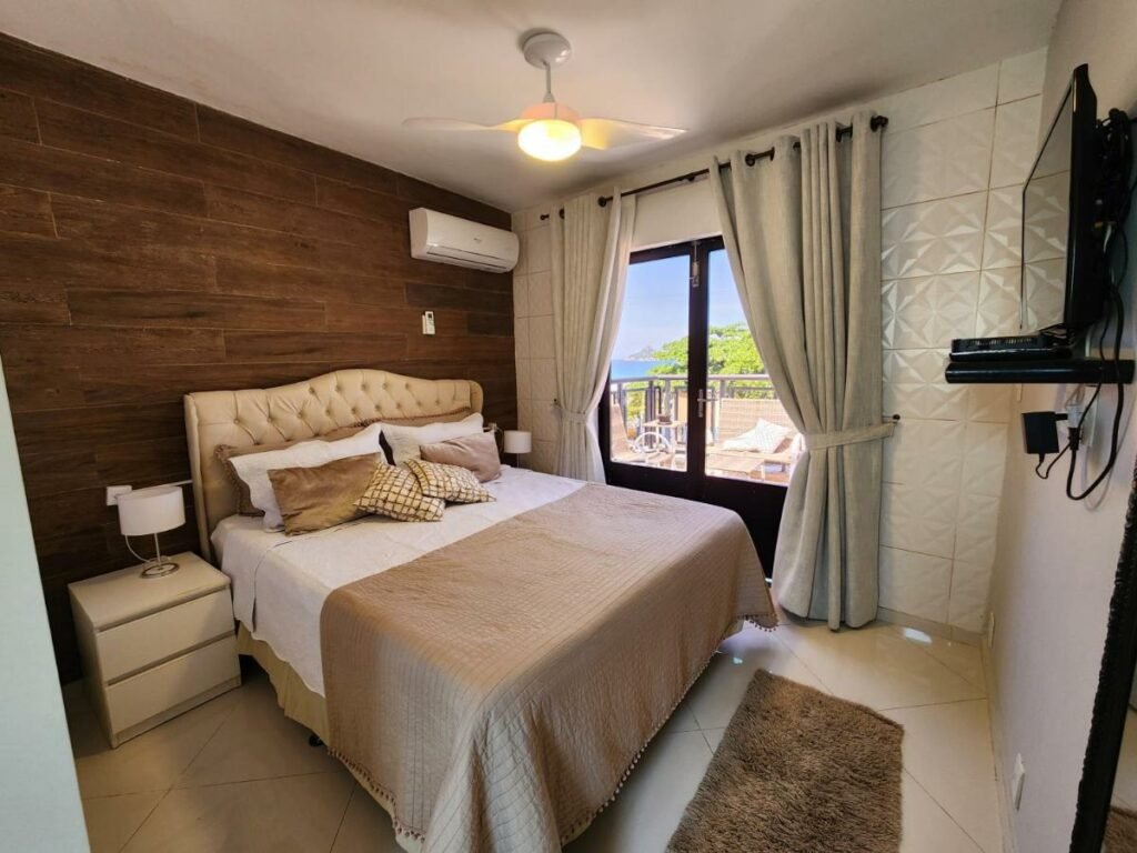 Quarto do AP Pepê ALECRIM com JACUZZI e SOLÁRIO Frente ao MAR com cama de casal do lado esquerdo da imagem com uma cômoda em cada lado da cama com luminária do lado esquerdo da cama uma porta que dá acesso a varanda.