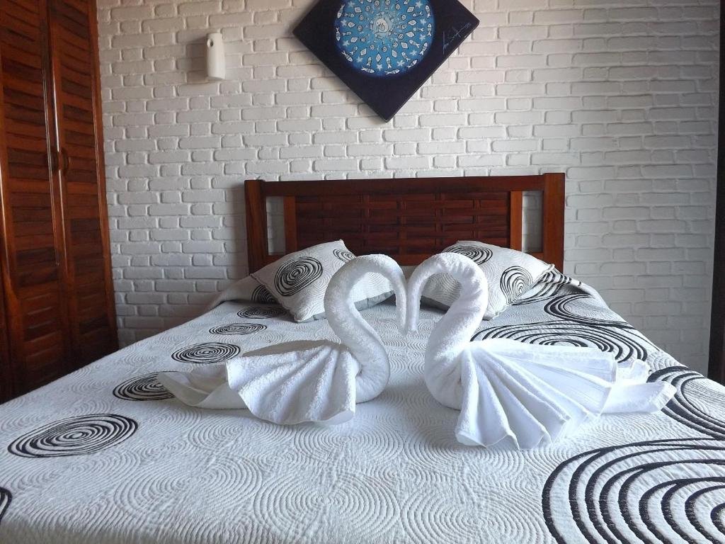 Quarto em Apartamentos Casa da Pedra, uma cama de casal, com toalhas dobradas em formato de cisnes, uma cabeceira de madeira, uma armário do lado esquerdo, um quadro sob a cama, parede de alvenaria branca de tijolinhos