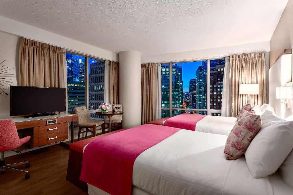 Quarto do Auberge Vancouver Hotel com janela panorâmica com vista para a cidade, há uma pequena mesinha com duas cadeiras perto de uma das janelas, ao lado, uma mesa de escritório com uma televisão e uma cadeira giratória, do lado direito, há duas camas de solteiro com travesseiros e almofadas, o chão é de madeira