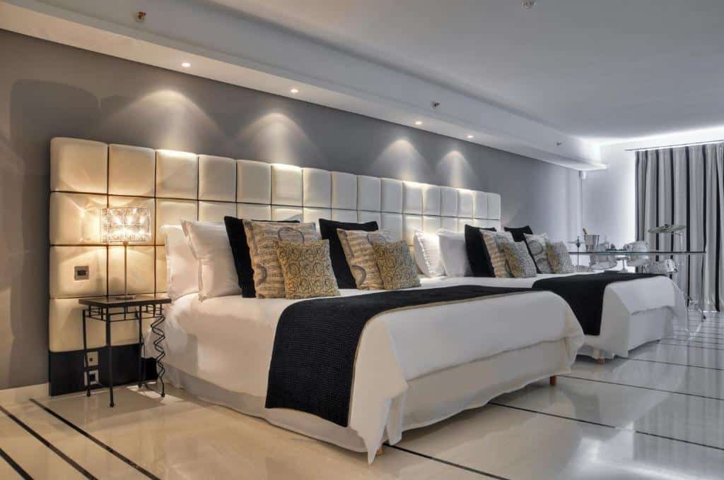 Quarto amplo no AX The Palace com duas camas de casal com muitos travesseiros e almofadas, tudo em tons de cinza e branco, há também iluminação indireta sob as camas e um abajur, para representar hotéis em Malta