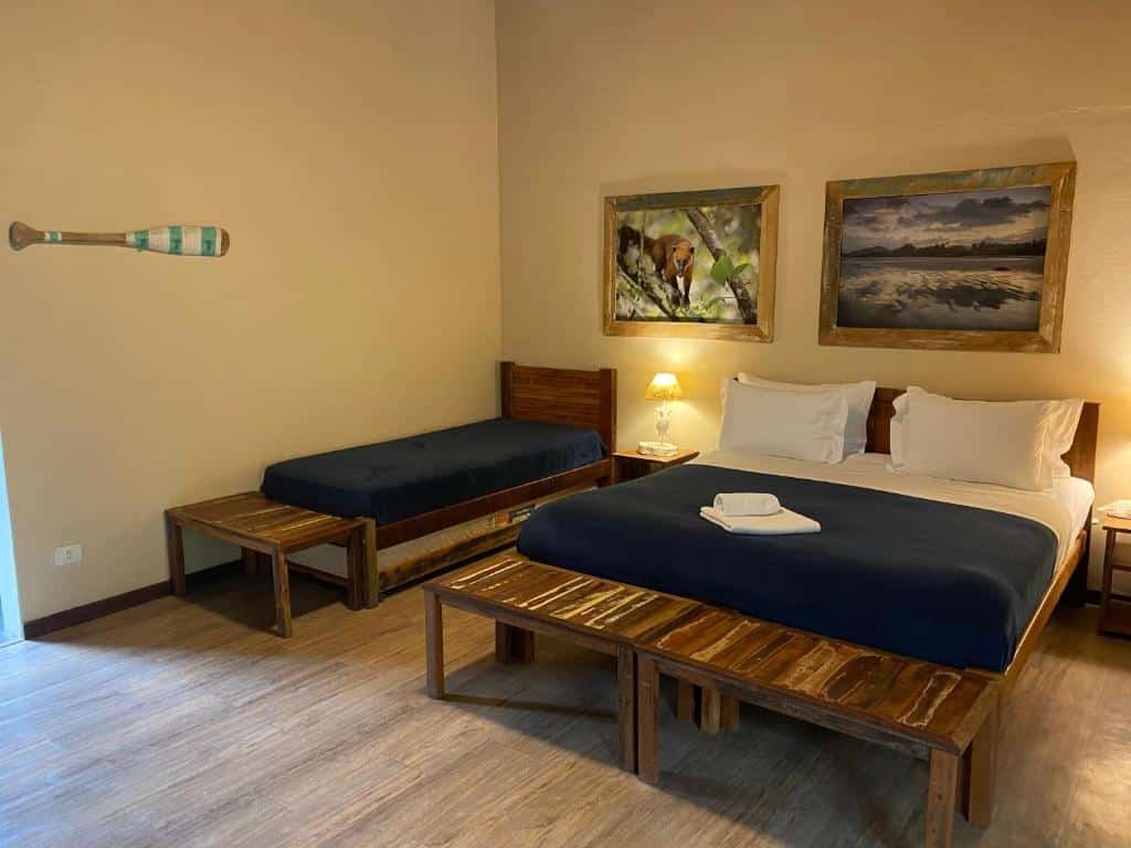 Quarto do Banana Bamboo Ecolodge com uma cama de casal e uma cama de solteiro. Nos pés de cada cama tem um banco de madeira e do lado de cada cama tem uma mesa de cabeceira. Além disso, no quarto tem dois quadros na parede e um chão de madeira.