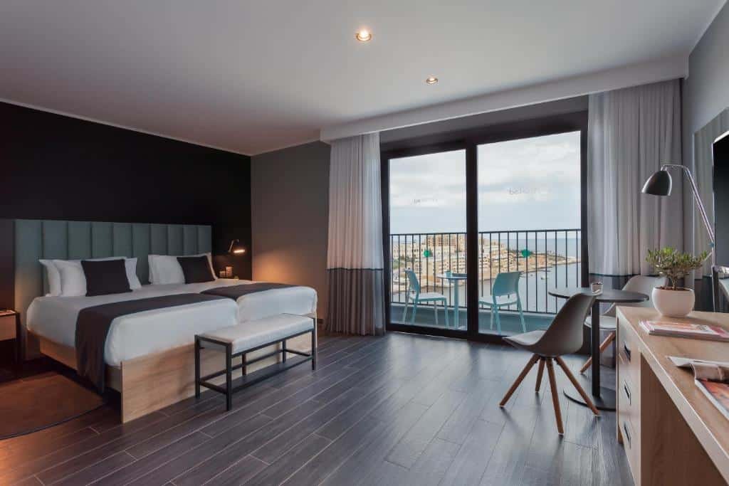 Quarto espaçosos no be.HOTEL com uma varanda com cortinas, duas camas de solteiro, uma mesa com duas cadeiras, uma televisão, tudo em tons de branco, cinza e marrom claro, o piso é de maneira, para representar hotéis em Malta