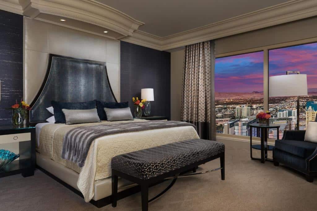 Quarto no Bellagio com uma janela panorâmica com vista para a cidade, ao lado da janela tem uma poltrona preta e uma pequena mesinha, do lado esquerdo tem uma cama de casal e há um carpete bege claro, o local é decorado em cinza e preto