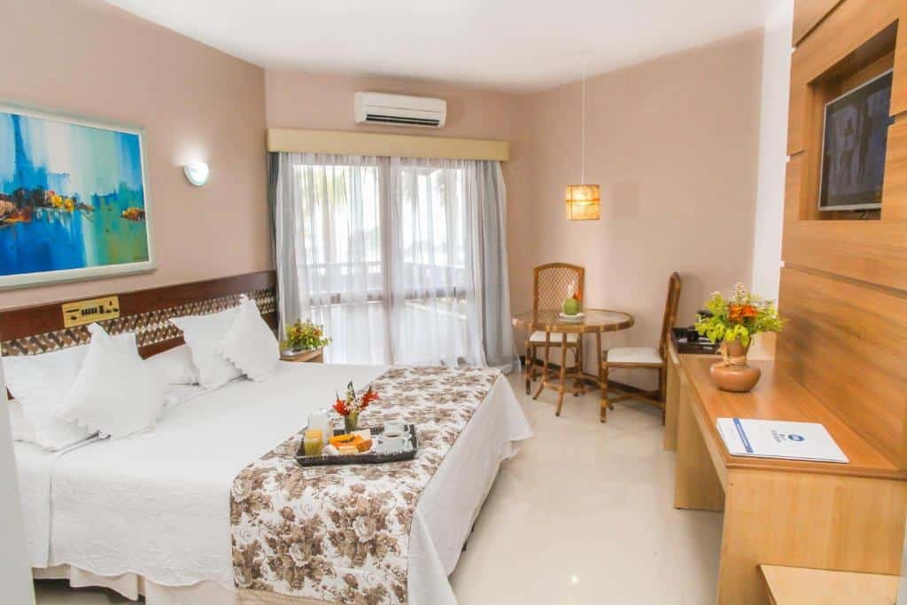 Suíte do Best Western Shalimar Praia Hotel. Há uma cama de casa na esquerda, com uma bandeja de café da manhã em cima. Na frente da cama há um painel de televisão com escrivaninha embutida. No fundo do quarto, mais à esquerda, há uma janela com varanda e uma mesa redonda com duas cadeiras ao lado.