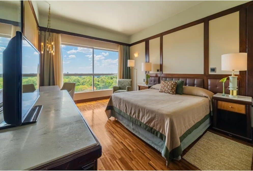 Quarto do Bourbon Cataratas do Iguaçu Resort com uma cama de casal e de cada lado da cama tem uma mesa de cabeceira e do lado direito tem uma poltrona e uma enorme janela de vidro. De frente pra cama tem uma mesa com cadeira e uma tv.