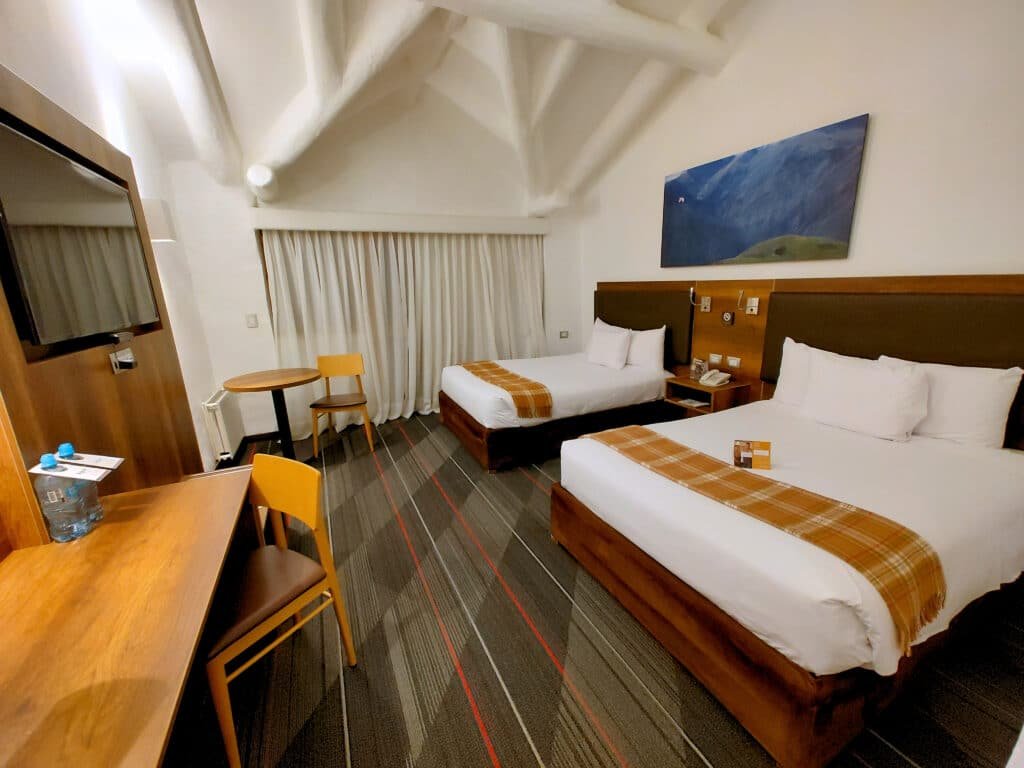 quarto duplo da Casa Andina Premium Valle Sagrado Hotel & Villas com duas camas de casal, lado a lado, na parte direita da imagem, O chão é acarpetado e em frente as camas há uma mesa de madeira para trabalhar, além de uma pequena mesa de madeira redonda para tomar café.