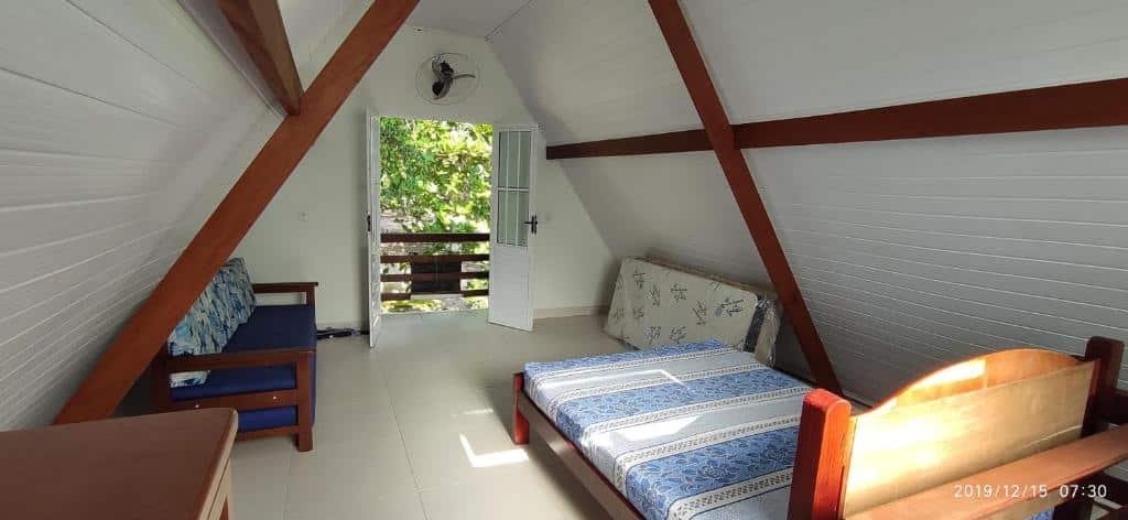 Quarto do Chale da Dalva com uma cama de casal, um colchão no chão do lado direito e do lado esquerdo uma mesa e um sofá. De frente com a cama tem uma porta aberta que dá acesso a varanda e um ventilador na parede em cima da porta.