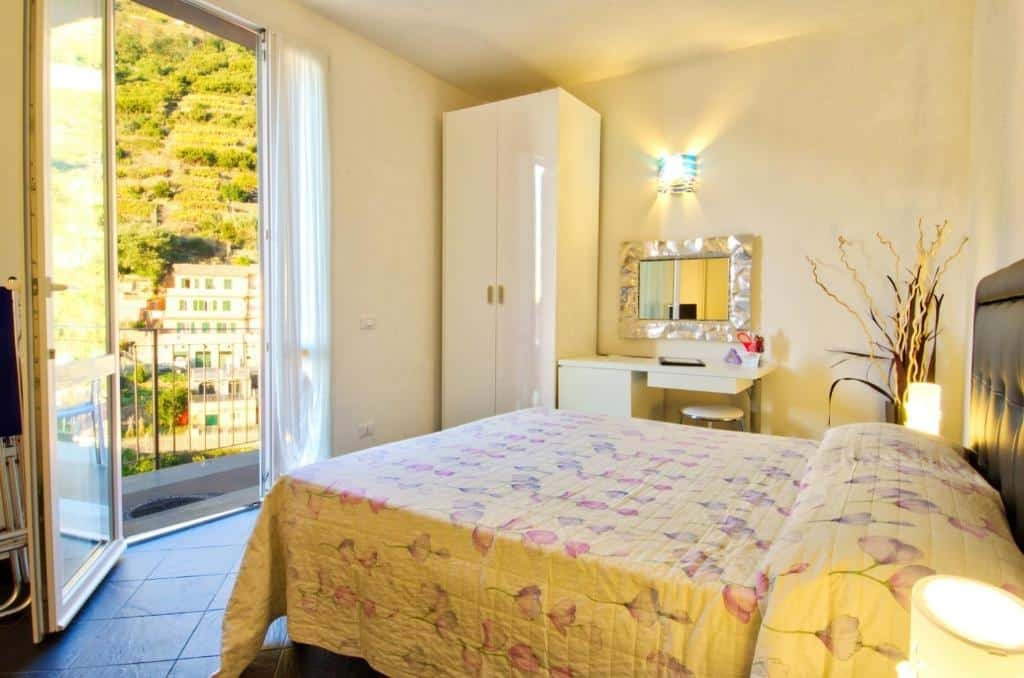 Quarto com uma cama de casal, penteadeira com espelho na parede, armário branco ao lado, uma planta de decoração e uma janela com sacada com vista para a cidade. 