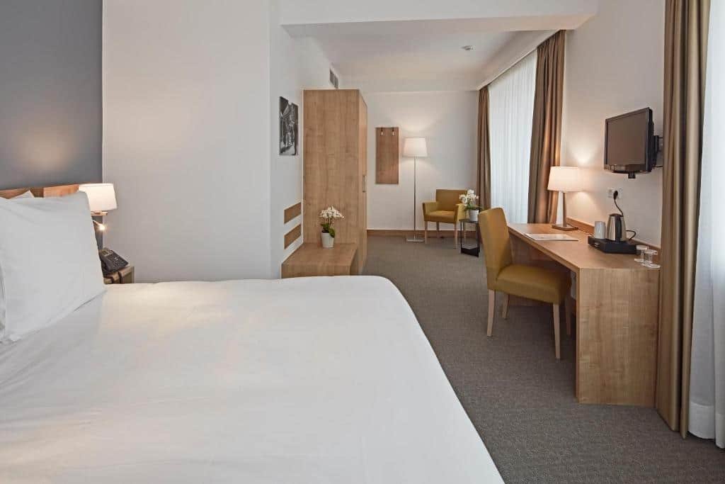 Suíte do City Hotel, para ilustrar o post sobre hotéis em Luxemburgo. A cama está no canto inferior esquerdo da foto. Ela é box e de casal. Ao seu lado direito notamos um abajur e um telefone. No fundo do quarto podemos ver uma escrivaninha com cadeira e amenidades para chá e abajur sobre sua superfície. Uma TV também está fixa acima da mesa. Atrás, há um guarda-roupa, e mais ao fundo, há uma poltrona entre um abajur de chão e uma mesa com flores em cima, ao lado de uma janela.