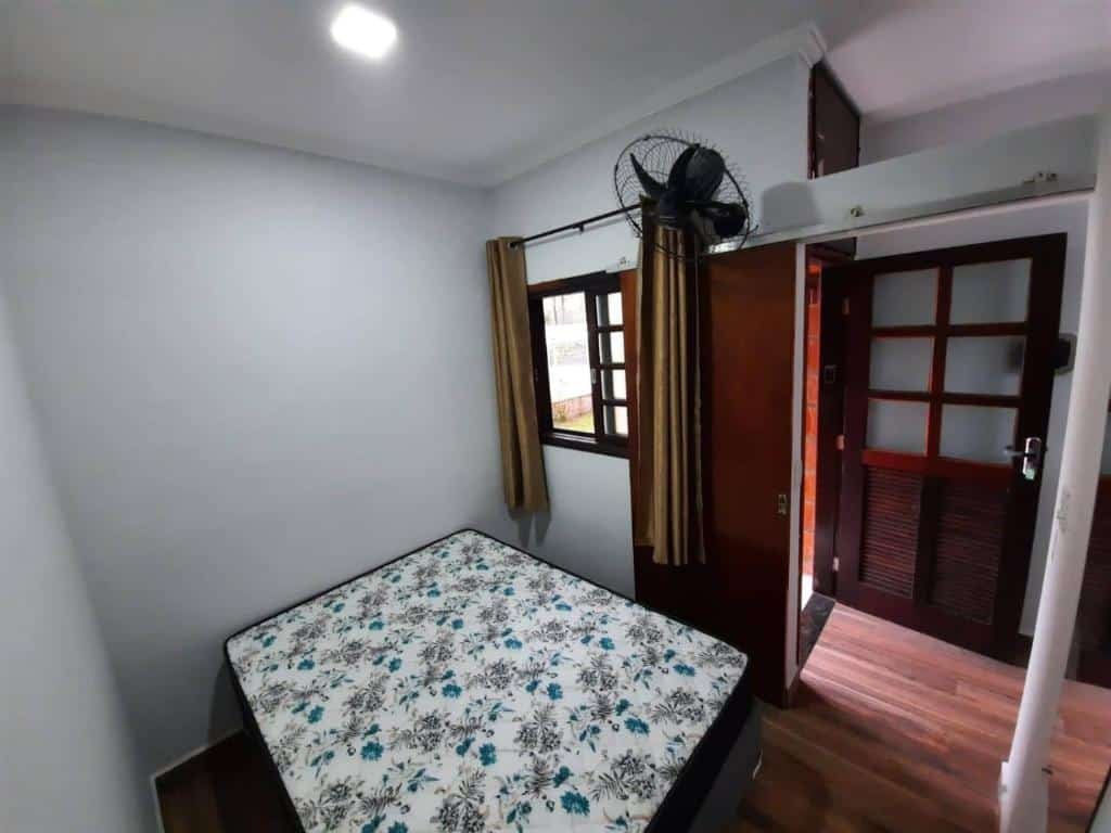 Quarto do Condomínio Ville I que mostra uma cama de casal com uma janela com cortinas e um ventilador na parede direita. Na frente da cama tem um espelho e do lado mais ao fundo tem uma porta que dá acesso a outro cômodo.