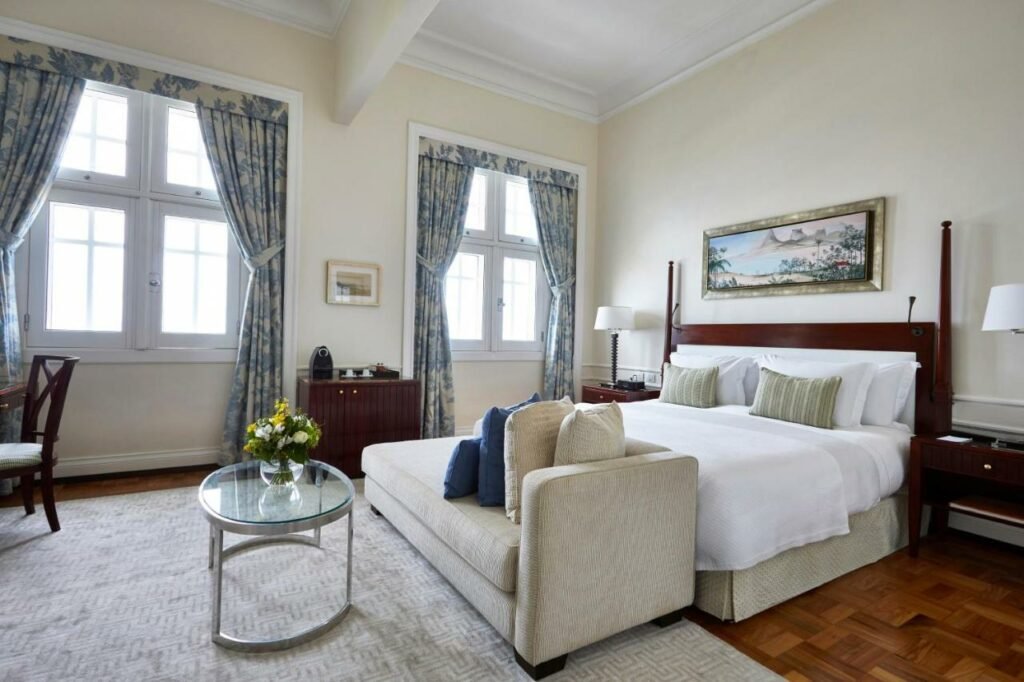 Quarto do Copacabana Palace com cama de casal do lado direito da imagem com uma cômoda em cada lado da cama com luminária, no pé da cama um sofá e em frente ao sofá uma mesa de centro.