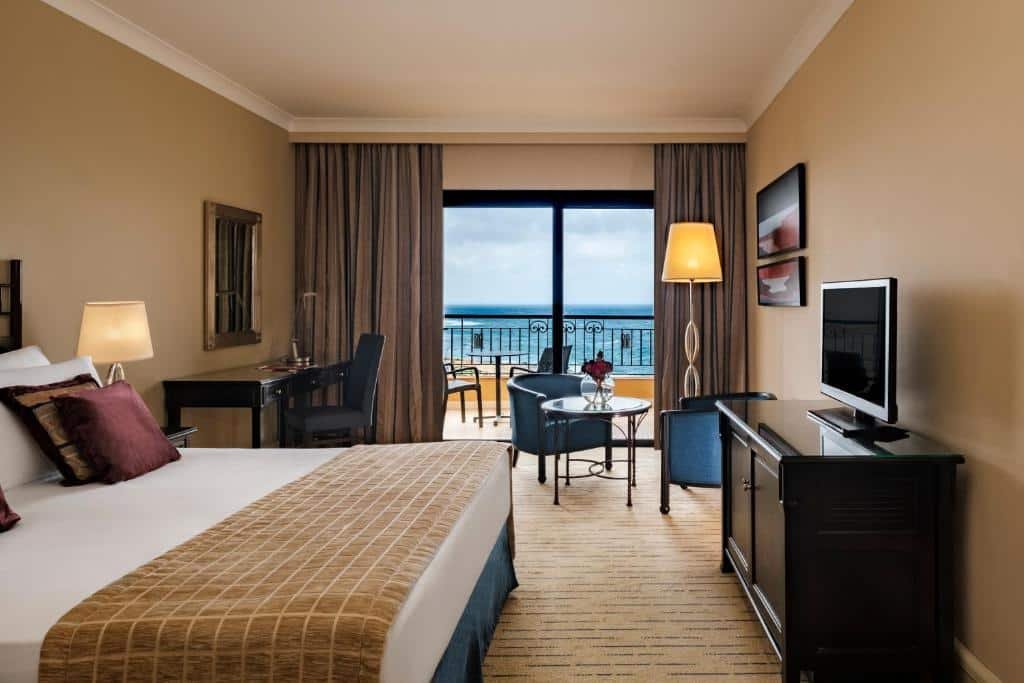 Quarto do Corinthia Hotel St. George’s Bay com uma sacada dando vista para o mar, uma cômoda com televisão, uma cama de casal, uma mesa de escritório com uma cadeira, duas poltronas e um abajur de chão