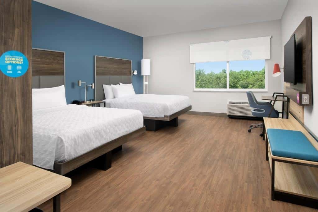 quarto amplo do Tru By Hilton Orlando Convention Center, um dos hotéis na International Drive em Orlando, com duas camas de casal, ambos com mesinha e luminária, há cadeira e balcão, há uma janela com veneziana