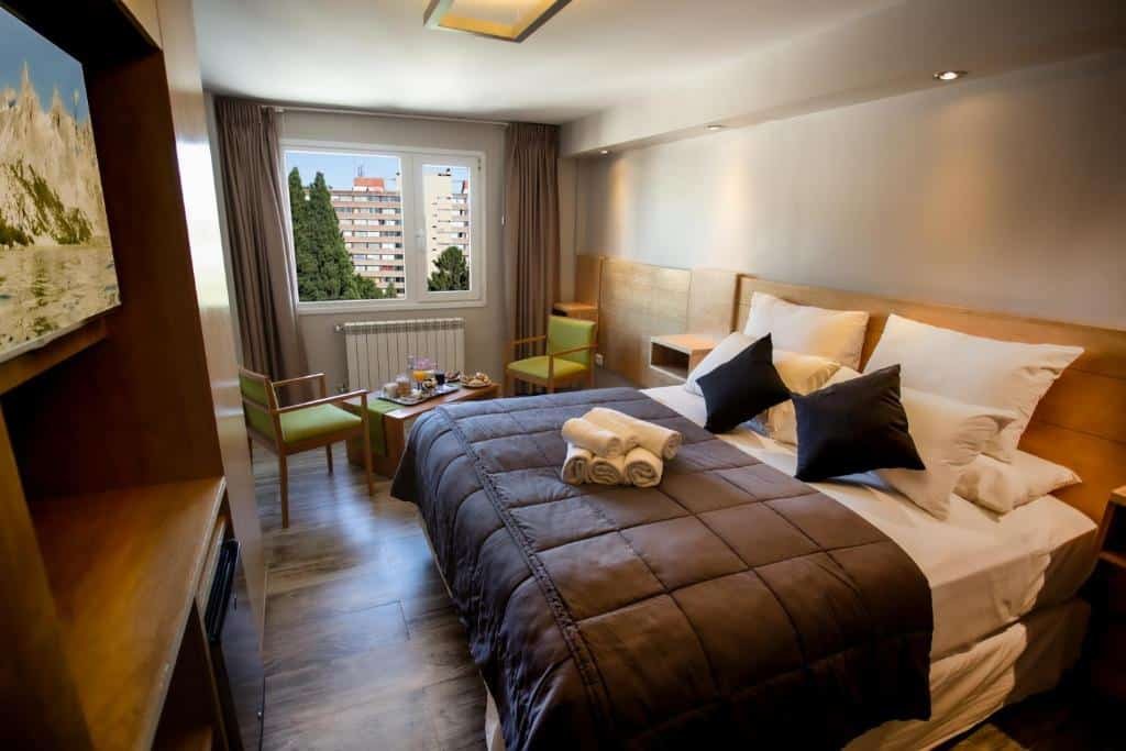 Quarto do Hotel EcoSki by bund com cama de casal, frigobar em frente a cama em uma cômoda com TV a cima, do lado esquerdo tem duas cadeiras e uma mesa em frente a uma janela com cortinas cinzas.