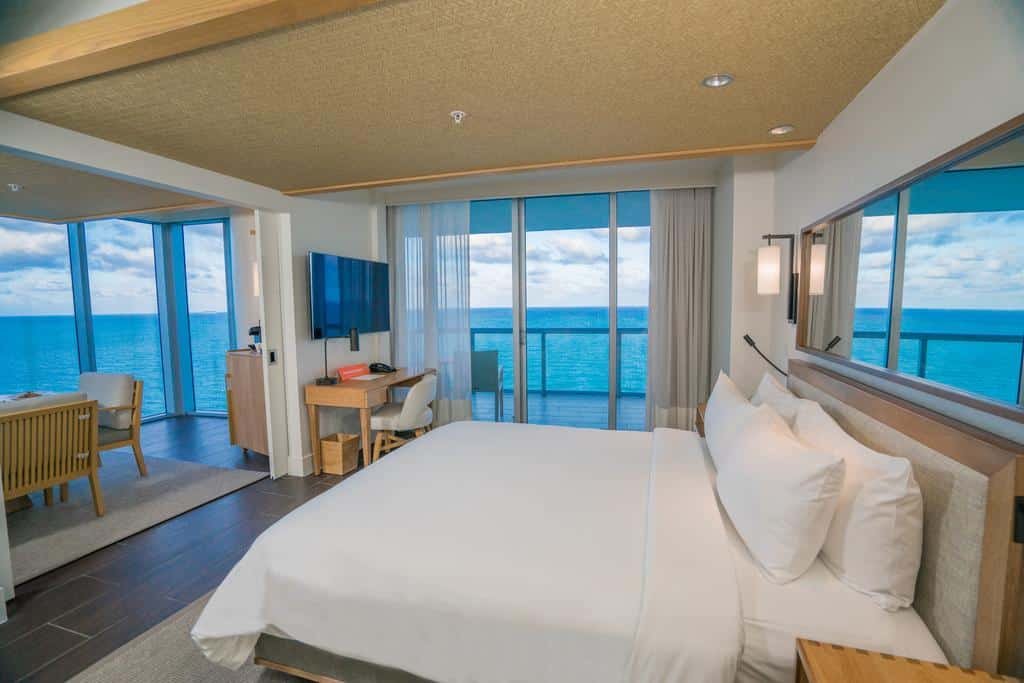 A imagem mostra um quarto moderno do Eden Roc Miami com uma grande janela de vidro que oferece uma vista deslumbrante do oceano. O quarto tem uma cama de casal grande com roupa de cama branca e almofadas macias. Há uma TV de tela plana montada na parede em frente à cama e uma pequena mesa de trabalho com cadeira. À esquerda, uma área de estar com cadeiras e uma mesa também oferece vista para o mar. O ambiente é iluminado naturalmente, com uma decoração minimalista e sofisticada, criando uma atmosfera relaxante e luxuosa.