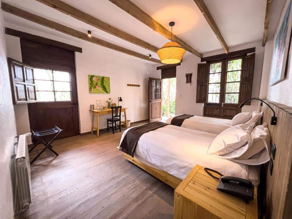 suíte do El Albergue Ollantaytambo com duas camas de solteiro no lado direito da imagem. Em frente as camas, há uma pequena mesa de madeira com uma cadeira, para trabalho. Há vários elementos decorativos em madeira, tanto no teto quanto nos móveis