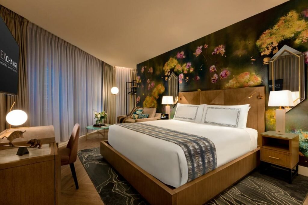 Quarto do EXchange Hotel Vancouver decoração com um papel de parede com rosas desenhadas atrás da cama de casal, duas mesinhas de cabeceira de madeira com abajures em cima delas, o chão é de madeira, do lado esquerdo tem uma ampla janela com uma poltrona, um abajur de chão e uma pequena mesinha de centro, de frente com a cama, há uma mesa de escritório com uma cadeira, e uma televisão presa na parede sob a mesa, para representar hotéis em Vancouver