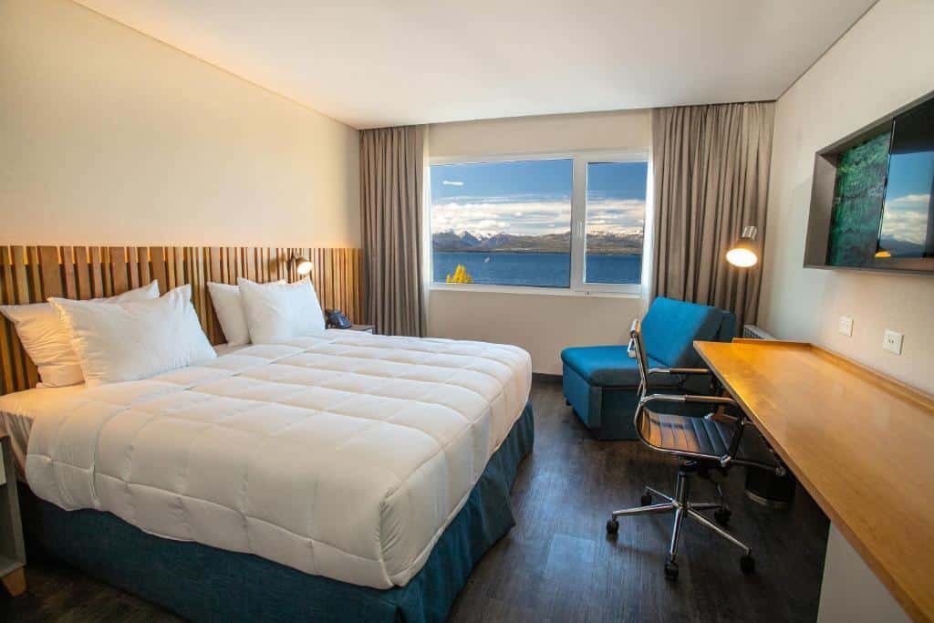 Quarto do Hampton By Hilton Bariloche com cama de casal, mesa do lado esquerdo, mesa de trabalho com cadeira em frente a cama, TV na parede em cima da mesa de trabalho, sofá cor azul do lado esquerdo e janela ampla do lado esquerdo com cortinas cinzas