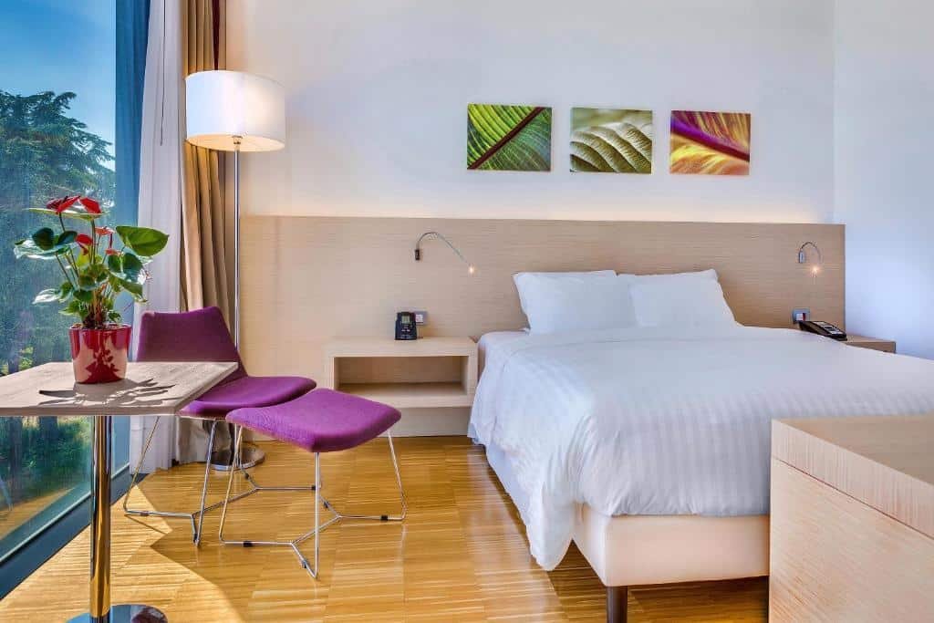 Quarto no Hilton Garden Inn Venice Mestre com uma cama de casal, em estilo mais moderno e atual, com uma poltrona roxa, alguns quadros na parede, chão de madeira e uma ampla janela, além de um abajur de chão e um vaso de flor