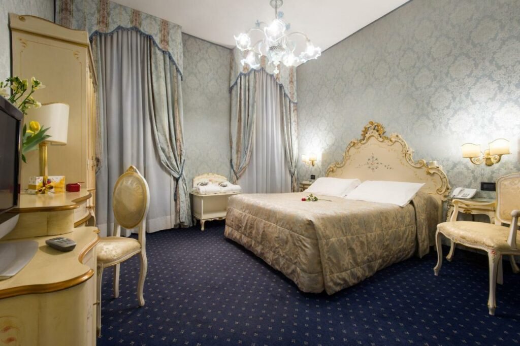 Quarto espaçoso no Hotel Carlton On The Grand Canal  em tons de azul escuro e claro, com detalhes em bege, estilo renascentista, cama de casal, duas poltronas, abajures nas paredes, uma televisão e duas janelas com cortinas